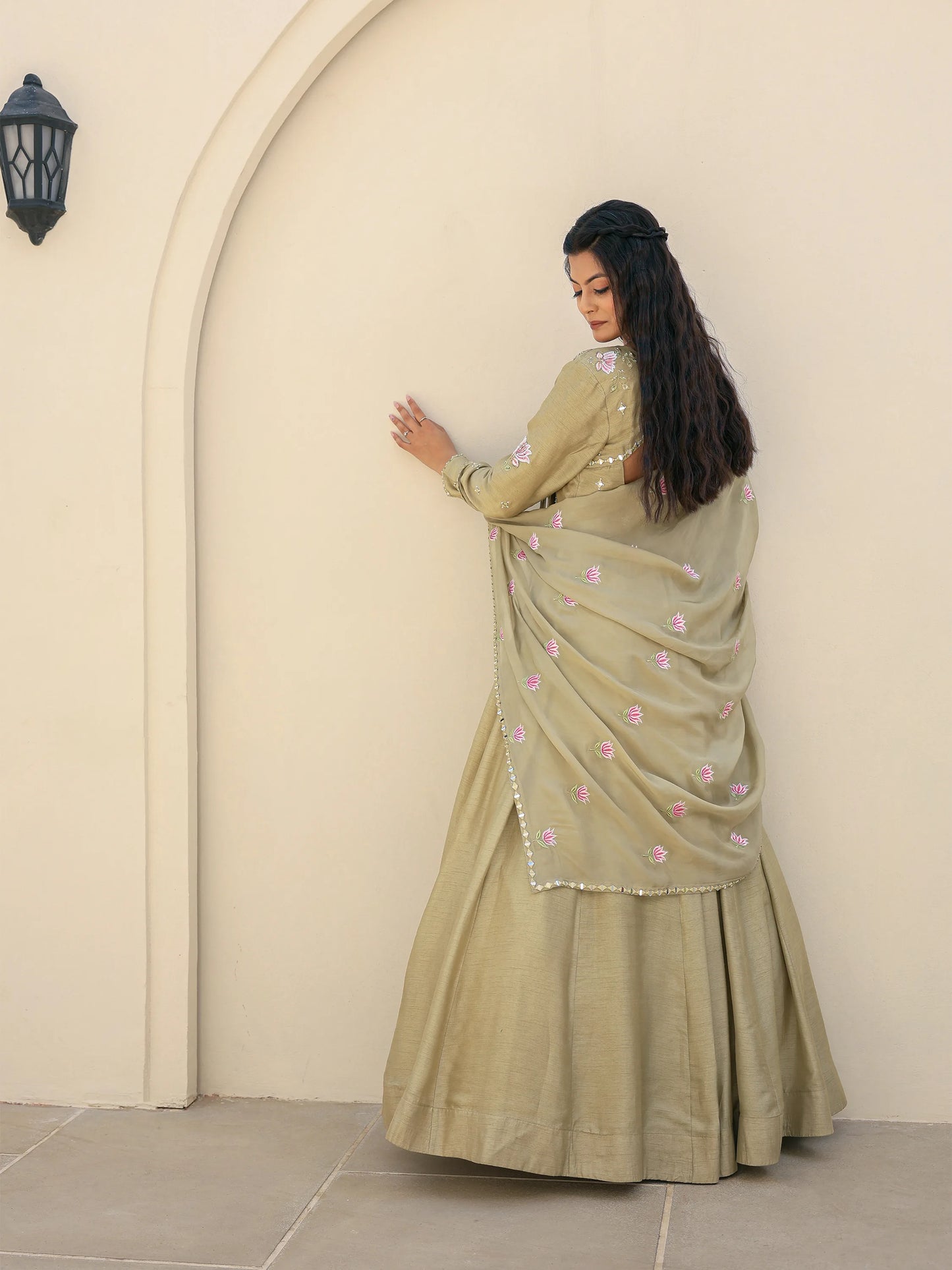 Woman wearing a sage green Anarkali with floral motifs on the dupatta standing against a beige wall.