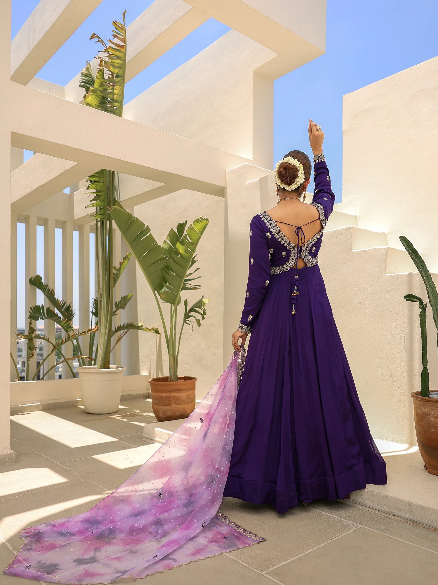Woman in a purple anarkali wearing a floral headband, standing on a rooftop with plants under a blue sky.