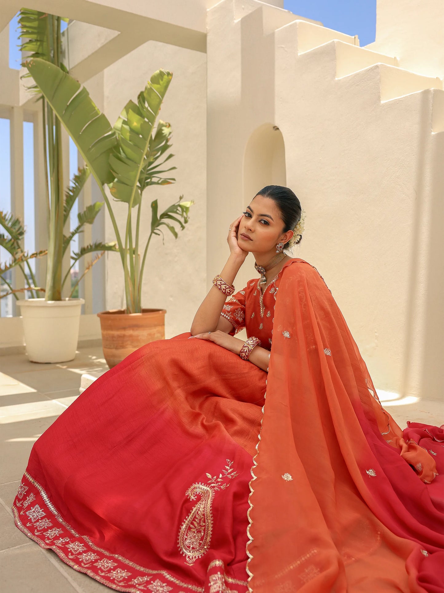 Model seated in a rustic orange Anarkali detailed with intricate zardosi work and a shaded dupatta.