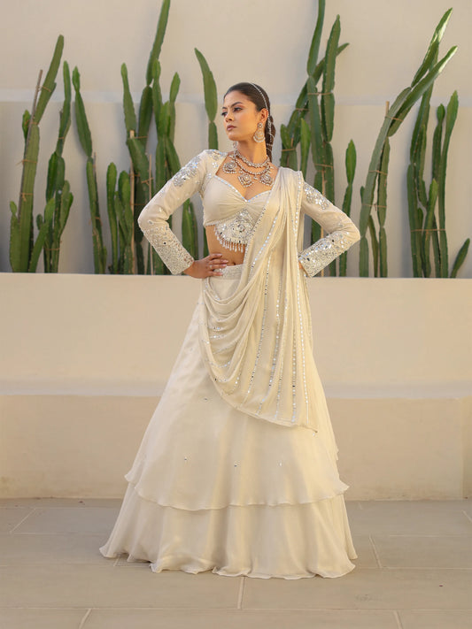 Woman wearing a white drape saree with a matching white blouse, standing against a neutral wall with green plants.