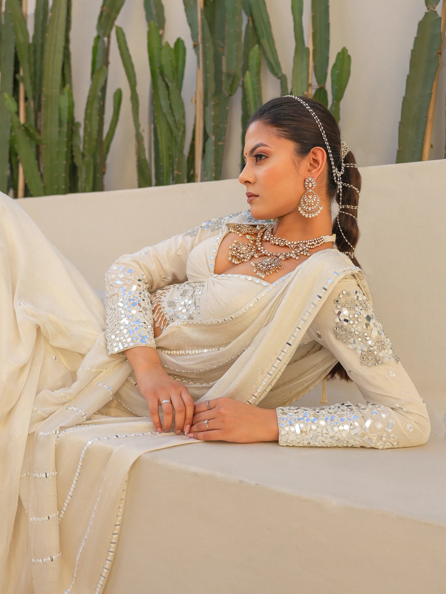 Woman wearing a white drape saree with jewelry, sitting on a wall with plants in the background.