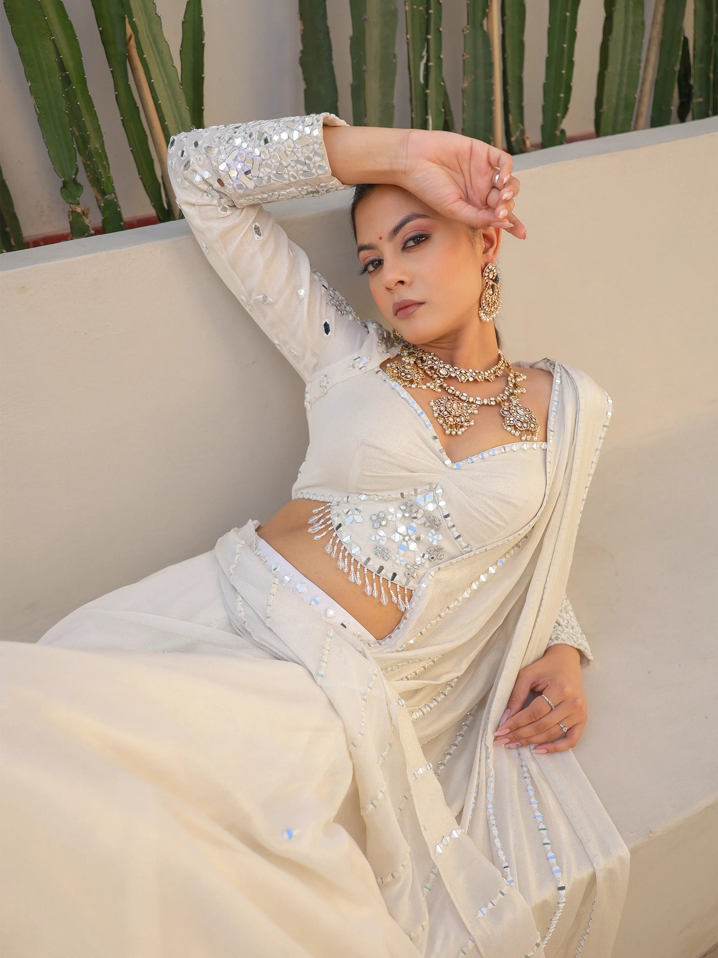 Woman wearing a white saree with mirror work, sitting on a wall.
