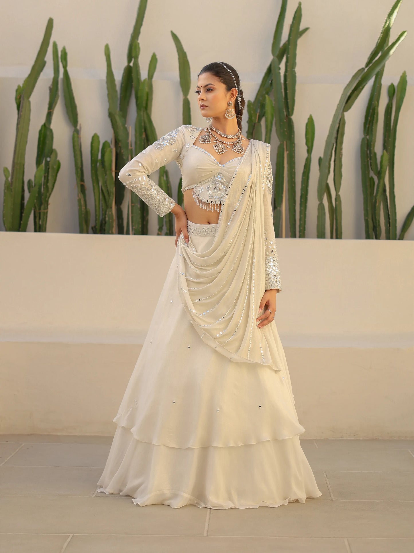 Woman wearing a white shimmer georgette drape saree with mirror work, standing against a neutral background with plants.