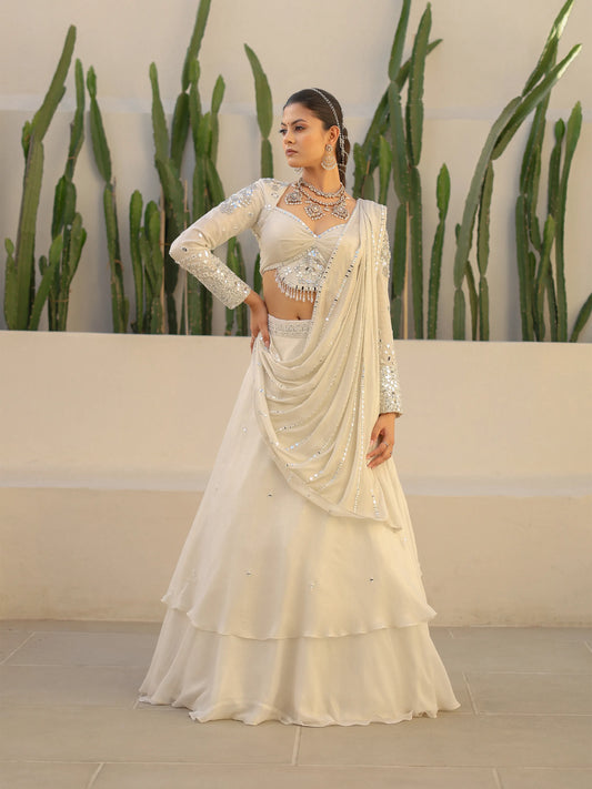 Woman wearing a white shimmer georgette drape saree with mirror work, standing against a neutral background with plants.